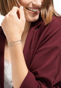 Bracciale d'argento con charm a forma di cuore e freccia, design a doppia catena, indossato su un polso, che completa una giacca texturizzata bordeaux.