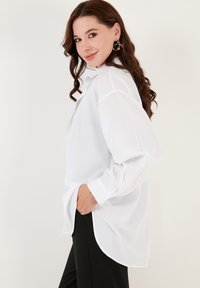 Hvid oversized skjorte med knapper lavet af letvægtsstof, med lange ærmer, en høj-lav kant og sidelommer.
