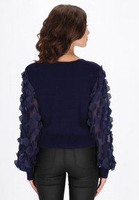 Laivastonsininen pusero, jossa on pitkät, läpikuultavat hihat, koristeltu kuusikulmaisilla röyhelöillä. Istuvan mallinen, jossa resorikoriste kaula-aukossa ja helmassa.