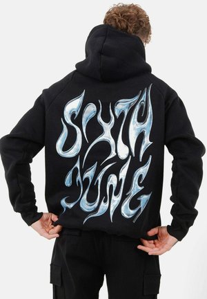 Schwarzer Hoodie mit großen, stilisierten weißen und blauen Buchstaben auf dem Rücken. Hergestellt aus dickem Stoff mit einer strukturierten Oberfläche.