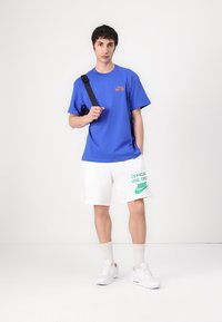 Mėlyna trumpomis rankovėmis marškinėliai su oranžiniu logotipu, derinami su baltais šortais, ant kurių yra žalias tekstas ir logotipas, užbaigta baltais sportbačiais.