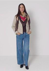 Beige kort jacka över en svart topp, parat med vida blå jeans och svarta stövlar. En flerfärgad halsduk ger kontrast runt halsen.