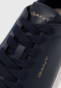 Zbliżenie granatowego buta sportowego z grubymi sznurowadłami i złotym logo GANT na języku oraz z boku.
