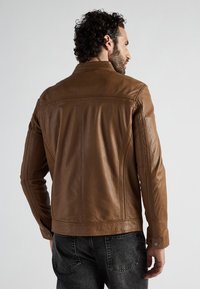 Mann mit lockigem Haar, der eine figurbetonte braune Lederjacke und schwarze gewaschene Jeans trägt, von hinten gegen einen hellen Hintergrund gerichtet.
