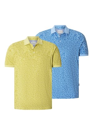 Mann trägt gelbes Polohemd mit Blumenmuster und Kragen sowie dunkelblaue Hose, steht mit einer Hand in der Tasche vor einfarbigem Hintergrund.