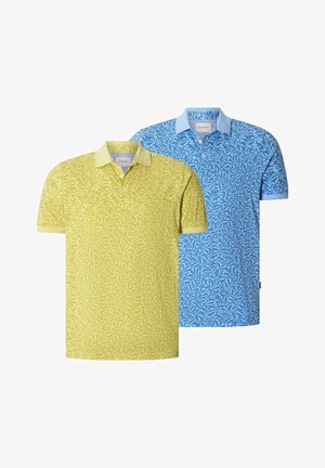 Mann trägt gelbes Polohemd mit Blumenmuster und Kragen sowie dunkelblaue Hose, steht mit einer Hand in der Tasche vor einfarbigem Hintergrund.