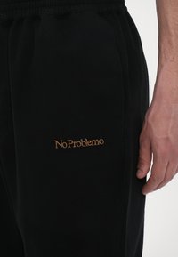 Black sweatpants made of soft fabric with an elastic waistband. Features "No Problemo" embroidered in gold on the left side.
