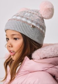 Graue Strickmütze mit pinkfarbenen Bommel, dekorativen Mustern und einem herzförmigen Metallakzent. Kombiniert mit einer pinkfarbenen Steppjacke mit Fellbesatz.