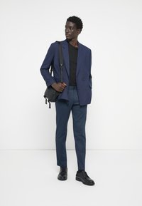Blazer bleu marine croisé sur un t-shirt noir, associé à un pantalon bleu foncé et des chaussures noires. Sac bandoulière noir avec des détails texturés.