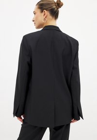 Blazer noir sur mesure avec un col classique, des manches longues et une coupe droite. Il présente une fente unique à l'arrière et une texture de tissu douce.