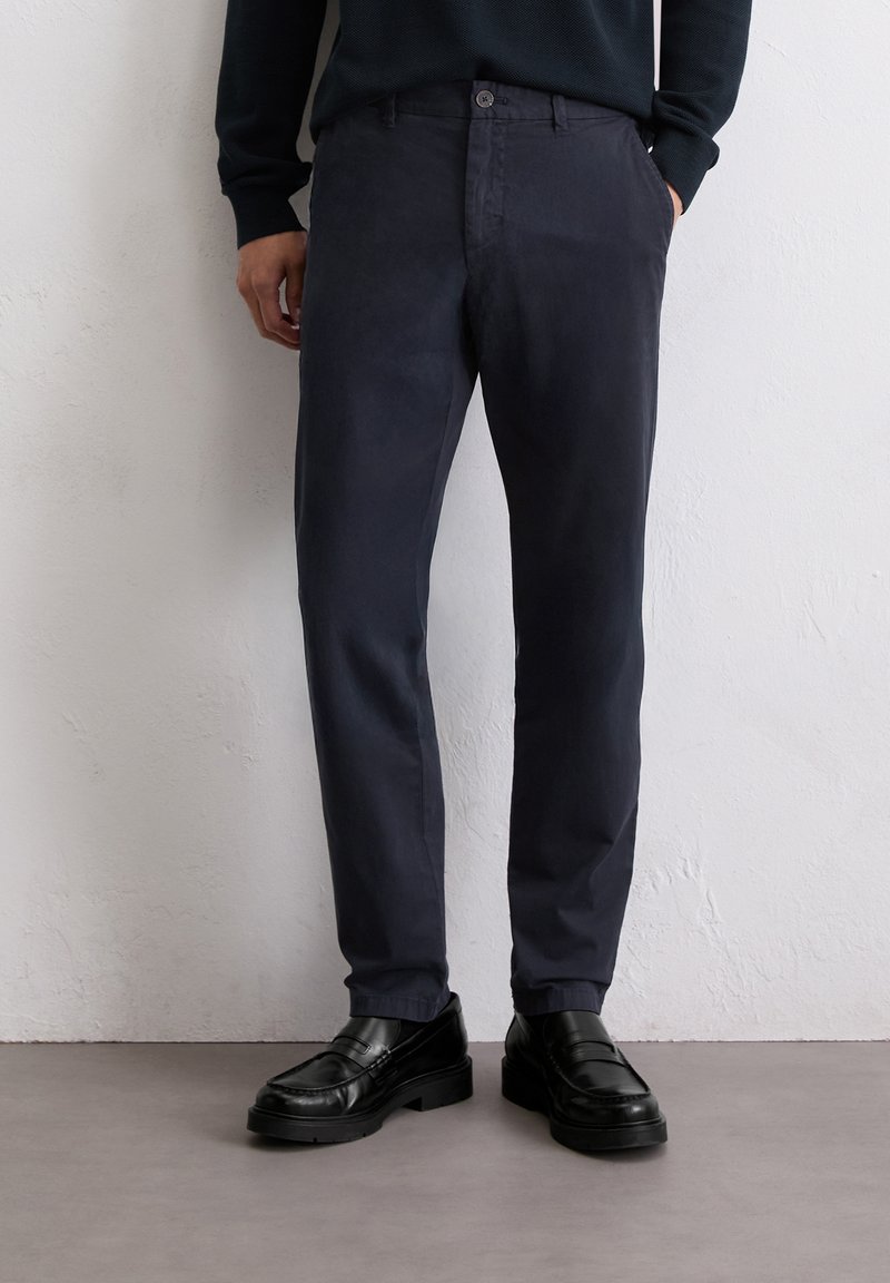 Marineblaue Baumwollmischungshosen mit geradem Schnitt, mit vorderen Taschen, einem Knopfverschluss und einer glatten Textur. Kombiniert mit schwarzen Slippern.