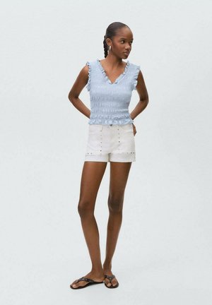 Top con scollo a smock di un bleu chiaro, con bordi arricciati, abbinato a pantaloni corti bianchi caratterizzati da dettagli con bottoni argentati e una vestibilità comoda. Sandali piatti completano il look.