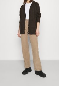 Hnědý pletený cardigan s ribkovanými okraji, vrstvený přes bílou košili. Spárováno s béžovými kordovými kalhotami s vysokým pasem a robustními černými botami.