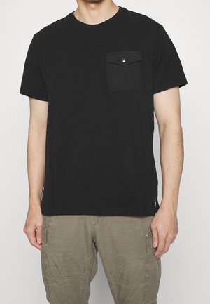 Mann trägt ein schwarzes T-Shirt mit kurzen Ärmeln, einer geknöpften Brusttasche und olivgrünen Cargohosen vor einfarbigem Hintergrund.