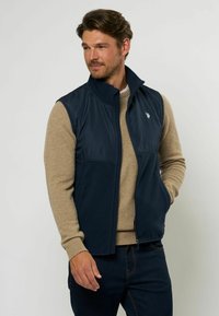 Marinblå väst med hög krage, tillverkad av ett slätt och lätt material. Har en fleecefoder och en subtil logotypdetalj på bröstet.