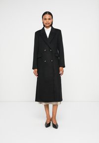Casaco preto de dois botões com lapelas recortadas, bolsos laterais e tecido texturizado, que se estende até a metade da panturrilha. Usado sobre um vestido de cor clara.