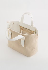 Sac fourre-tout en toile beige avec des poignées en cuir blanc et des finitions en or, doté d'une poche zippée intérieure et d'un design structuré.