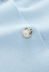 Jasnoniebieska tkanina o miękkiej, teksturowanej powierzchni oraz błyszczący srebrny guzik z falistym wzorem.