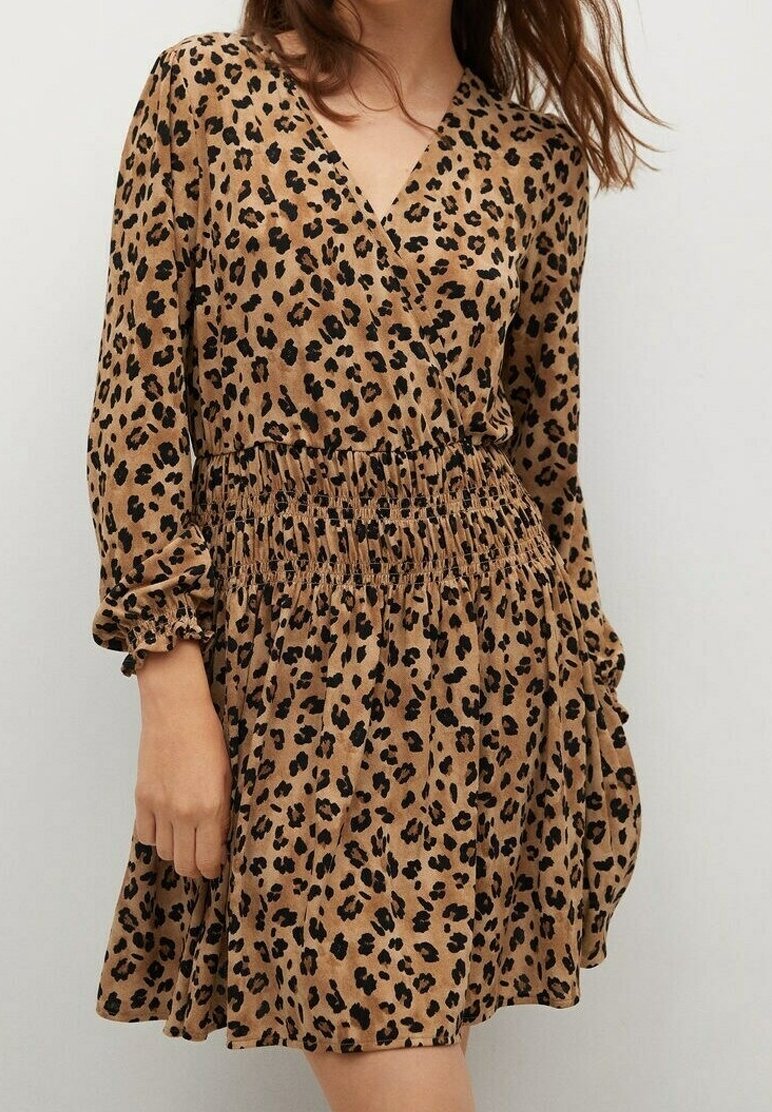 Femme portant une robe marron à motif léopard avec des manches longues et une taille froncée, debout contre un mur clair uni.