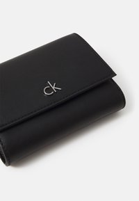 Juodas odinis piniginė su tekstūruotu paviršiumi, turintis sidabrinį CK logotipą priekyje ir sulankstomą dizainą. Kompaktiška ir paprasta.