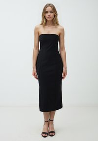 Schwarzes, trägerloses Midi-Kleid mit taillierter Silhouette, vertikalen Nähten und glattem Stoff. Kombiniert mit schwarzen Sandalen mit Knöchelriemen.