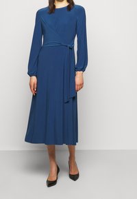 Robe midi bleu à manches longues avec un design enroulé et une taille nouée. Fabriquée en tissu doux, elle présente un col rond et une silhouette fluide.