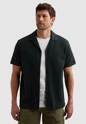 Mann mit schwarzem kurzärmligem Hemd mit Knöpfen über weißem T-Shirt und olivgrünen Cargohosen, schaut nach links auf einfarbigem Hintergrund.