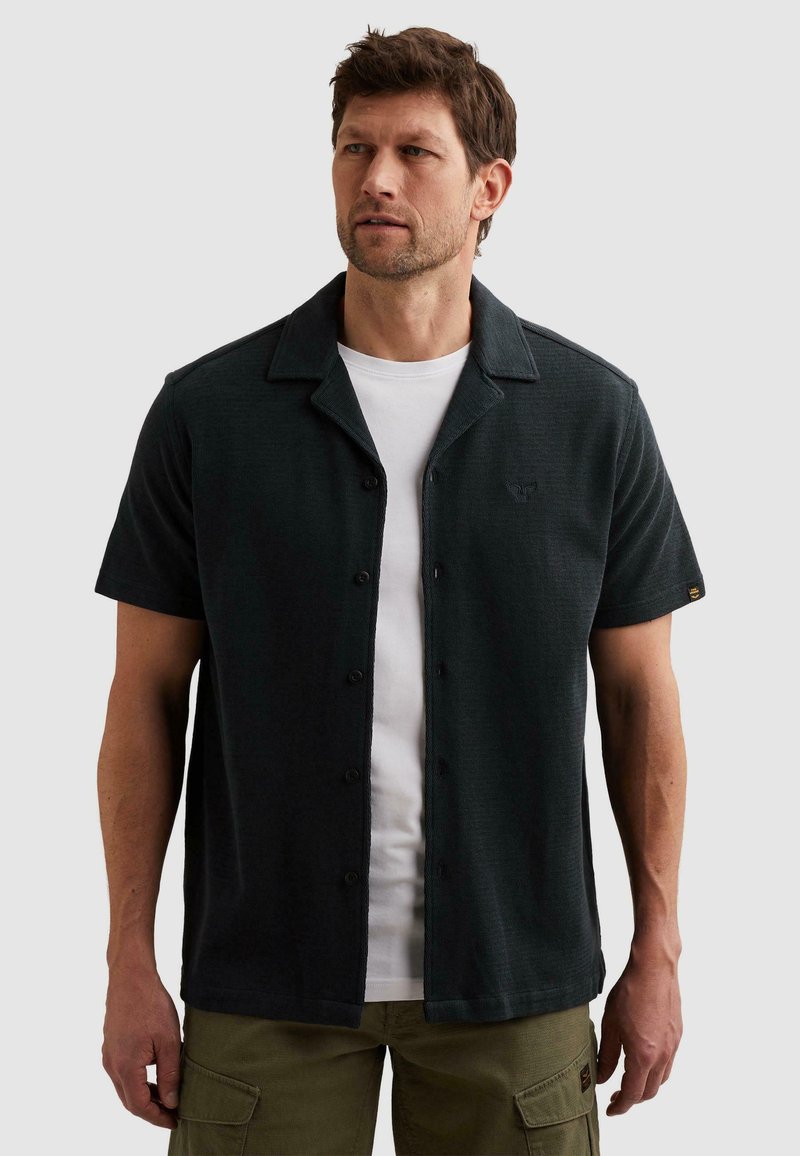 Homme portant une chemise noire à manches courtes boutonnée sur un t-shirt blanc et un pantalon cargo vert olive, regardant vers la gauche sur un fond uni.