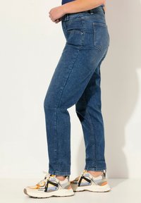 Modré džínové kalhoty se střihem rovného nohavice, obsahující zadní kapsy a zipové zapínání. V kombinaci s vícebarevnými sportovními teniskami.