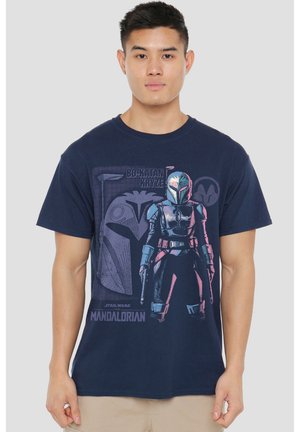 Young man wearing navy blue t-shirt with Bo-Katan Kryze character and Star Wars The Mandalorian logo on front against a gray background.