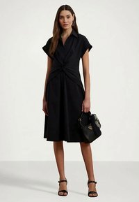 Jauna sieviete melnā ceļgala garuma kleitā ar sarullētām piedurknēm, turot melnā krāsā rokassomu un ģērbusies melnās sandalēs ar atklātu pirkstu.