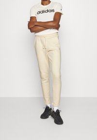 Camiseta blanca de Adidas con logo negro, joggers beige con cintura elástica, y zapatillas negras con calcetines blancos, de pie contra un fondo neutro.
