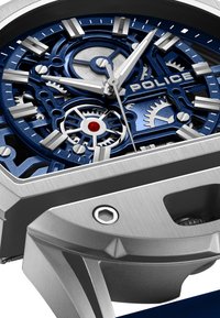 Srebrno in modro mehanično uro s odkrito zobniki, srebrnimi oznakami ur in imenom blagovne znamke "POLICE" na številčnici.