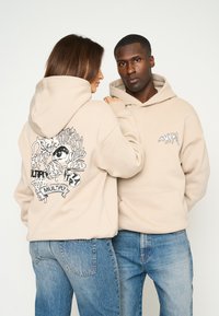 Sweatshirts beige avec un design graphique au dos en encre noire et un petit logo à l'avant. Coupe décontractée avec poches kangourou.