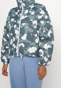 Personne portant une doudoune bleue et blanche à motif géométrique avec une fermeture éclair à l'avant et un pantalon bleu clair, debout devant un fond uni.