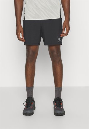 Mężczyzna ubrany w czarne sportowe spodenki z logo Odlo, szare skarpety za kostkę oraz czarne buty do biegania po szlakach z pomarańczowymi akcentami, stojący na białym tle.