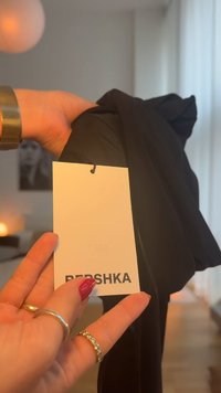 Svart plagg hålls i en hand och visar en vit lapp med texten "BERSHKA" i fetstil. Tyget verkar mjukt med en slät yta.