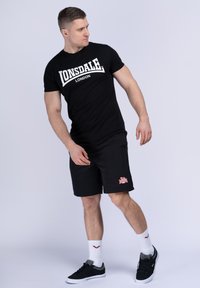 Zwarte Lonsdale t-shirt met wit logo, zwarte shorts met rood logo, zwarte schoenen en witte sokken met rode accenten. Neutrale achtergrond.