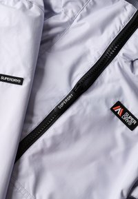 Jasnoszara, teksturowana kurtka z tkaniny z centralnym zamkiem błyskawicznym zakończonym czarnym suwakiem oraz naszywkami z logo w kolorach czarnym i czerwonym.