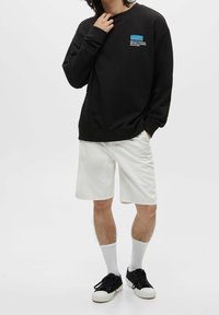 Czarna bluza z okrągłym dekoltem i ściągaczami przy rękawach, ozdobiona małą niebiesko-białą grafiką. Do tego białe szorty i czarne sneakersy.