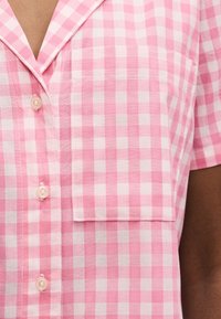 Πουκάμισο κοντομάνικο καρό ροζ και λευκό με μπροστινή τσέπη και κουμπιά, φορεμένο σε μεσαίο καφέ τόνο δέρματος.