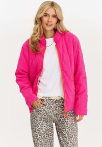 Jasnoróżowa pikowana kurtka z stojącym kołnierzem, zamkiem z przodu i pomarańczowymi akcentami, w połączeniu z dżinsami w panterkę.