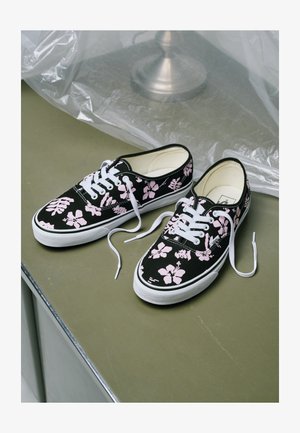 Sorte lave sneakers med hvide såler og pink blomsterprint, hvide snørebånd, der er løse, placeret på en olivengrøn overflade nær plastfolie.