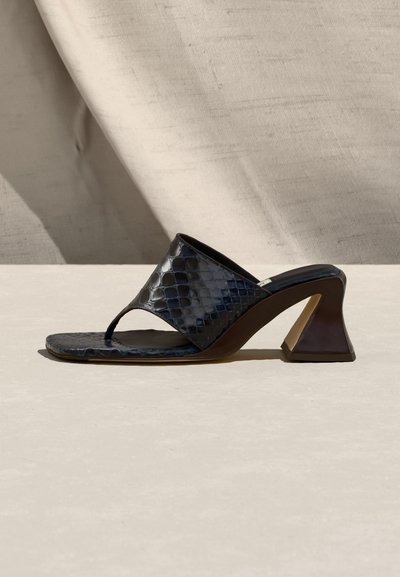 Μαύρο mule πέδιλο με ανοιχτά δάχτυλα, σχέδιο κροκόδειλου, τετράγωνη μύτη και χοντρό τακούνι πάνω σε μια μπεζ υφασμάτινη επιφάνεια.