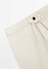 Gros plan sur un pantalon en lin beige avec un bouton noir, des passants de ceinture, des poches avant et une taille à lien noué.