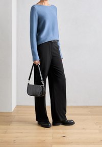 Maglione blu a maglia con maniche lunghe, abbinato a pantaloni neri ampi. Una piccola borsa nera con motivo coccodrillo è tenuta in una mano.