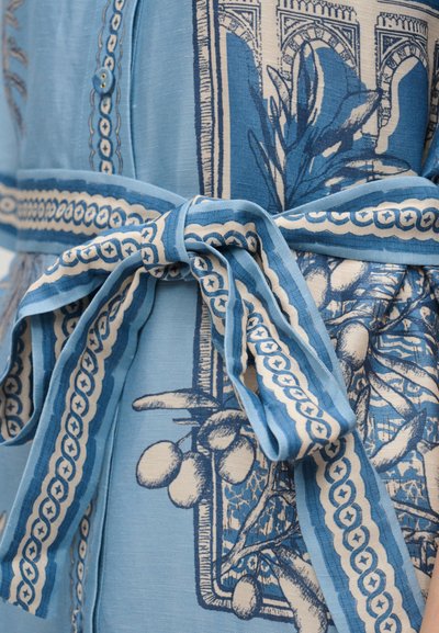 Μπλε υφασμάτινη ζώνη δεμένη σε φιόγκο γύρω από ένα φόρεμα με μπλε και κρεμ χρώμα, διακοσμημένο με μοτίβα από φυτά και αρχιτεκτονικές λεπτομέρειες.