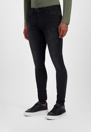 Mężczyzna w czarnych obcisłych dżinsach ze śladami lekkiego przetarcia i czarnych trampkach z białą podeszwą, stojący na jednolitym tle.