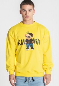 Sudadera amarilla con un gráfico de un oso de dibujos animados que lleva gafas y una gorra, con el texto "KAVAGH" debajo del diseño.