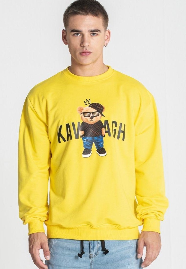 Sudadera amarilla con un gráfico de un oso de dibujos animados que lleva gafas y una gorra, con el texto "KAVAGH" debajo del diseño.